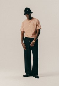 Muž s béžovým rebrovaným tričkom, čiernymi širokými nohavicami, čiernym klobúkom typu bucket, okuliarmi, zlatými hodinkami a náhrdelníkom, stojaci na jednofarebnom pozadí.