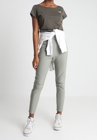 Khaki tričko s krátkým rukávem s logem, bílé mikina uvázaná v pase, slim-fit světle zelené kalhoty a bílé tenisky. Hladké textury.