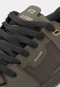 Green and brown synthetic sneaker with black laces, featuring embossed "GLOBE" logo, padded tongue, and textured ridges on the side panels.