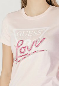 Camiseta de algodón de color rosa claro con cuello redondo, que presenta un logo triangular blanco y la palabra "amor" en strass rosa y plateado en la parte frontal.
