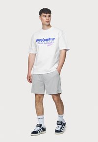Hvit t-skjorte med lilla og rosa grafisk tekst, grå shorts, svart og hvite joggesko, hvite sokker med logo, stående mot en ensfarget bakgrunn.