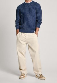 Navy Kabelstrickpullover mit Rundhalsausschnitt, kombiniert mit cremefarbenen, plissierten Hosen und beigen Sneakers mit hellbraunen Akzenten.