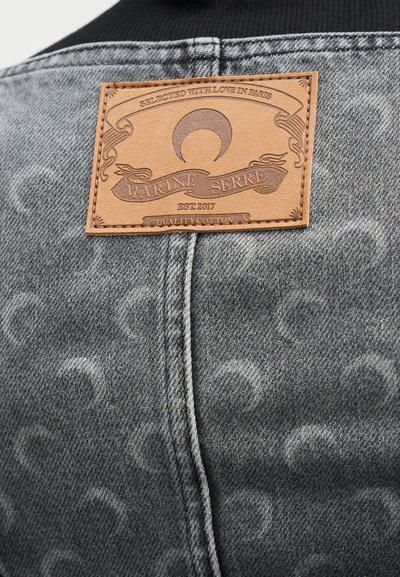 Γκρι τζιν ύφασμα με διακριτικό μοτίβο. Μια ταμπέλα από ταμπά δέρμα με ανάγλυφο κείμενο και σχέδιο είναι ραμμένη στο πίσω μέρος.