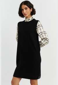 Schwarzes gestricktes ärmelloses Kleid mit geripptem Ausschnitt und seitlichen Schlitzen, getragen über einer weißen Bluse mit einem schwarzen Herzmuster.