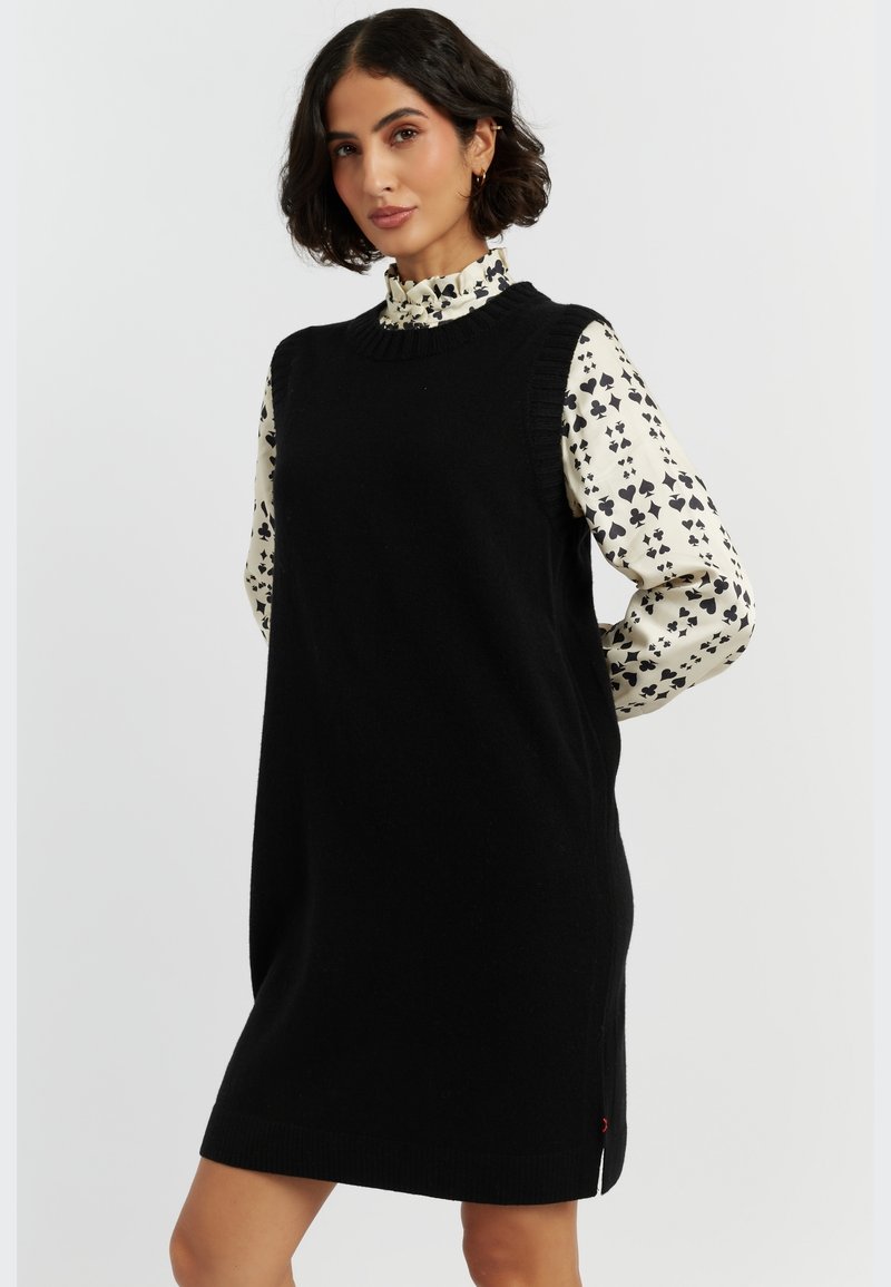 Schwarzes gestricktes ärmelloses Kleid mit geripptem Ausschnitt und seitlichen Schlitzen, getragen über einer weißen Bluse mit einem schwarzen Herzmuster.