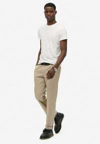 Camiseta blanca de cuello redondo, pantalones beige, zapatos de cuero negros con suela marrón. Reloj en la muñeca, pose de pie, fondo neutro.