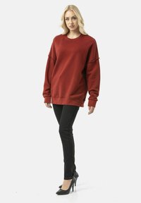 Rode sweatshirt met oversized pasvorm, geribbelde manchetten en verweerde accenten, gecombineerd met strakke zwarte jeans en zwarte hoge hakken.