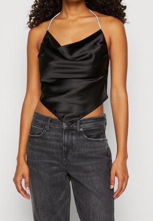 Kvinde iført sort satin haltertop med spids kant og mørkegrå højtaljede jeans mod hvid baggrund.