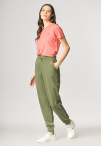 T-shirt corallo con un piccolo logo, abbinata a pantaloni jogger verde oliva con tasche laterali e polsini elastici, indossata con sneaker bianche.