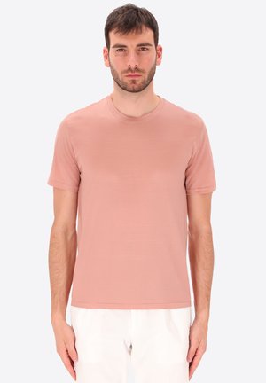 Uomo che indossa una semplice t-shirt rosa polvere a maniche corte e pantaloni bianchi, in piedi davanti a uno sfondo chiaro semplice.