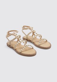 Sandalia con tiras que presenta correas de color beige claro adornadas con tachuelas metálicas doradas, suela plana y diseño de punta abierta.