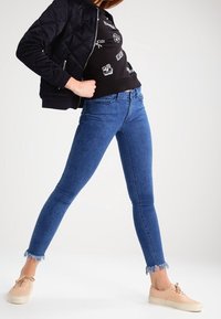 Vaqueros ajustados de mezclilla azul con dobladillo deshilachado, combinados con una sudadera negra con estampado gráfico y zapatillas de color beige. La chaqueta es acolchada y negra.