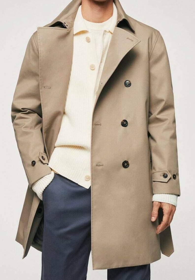 Homme portant un trench-coat beige sur un cardigan crème et un pantalon bleu foncé, avec une main dans la poche et l'autre détendue sur le côté.