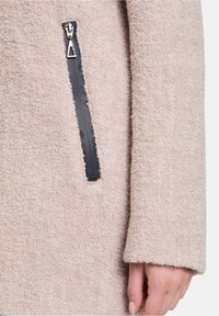 Un cappotto beige con trama, realizzato in tessuto morbido e con una tasca frontale nera con cerniera, caratterizzato da una silhouette rilassata e maniche lunghe.