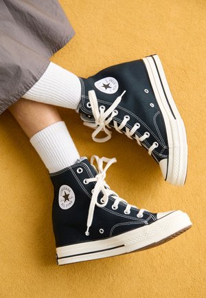 Beine übereinandergeschlagen mit schwarzen High-Top Converse All Star Sneakers, weißen Schnürsenkeln und weißen Rippsocken auf einem gelben Teppich.