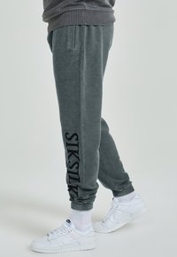 Grijze sweatpants van een gestructureerde stof, met een tapse pasvorm en een groot zwart logo op het linkerbeen, gecombineerd met witte sneakers.