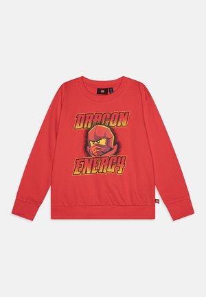 Κόκκινη υφαντή μπλούζα με γραφικό σχέδιο ενός θωρακισμένου χαρακτήρα και το κείμενο "ΔΡΑΚΟΝΤΙΑ ΕΝΕΡΓΕΙΑ" σε έντονα κίτρινα και πορτοκαλί γράμματα. Απαλή ύφανση, στρογγυλή λαιμόκοψη.