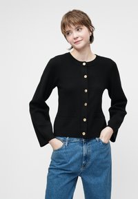 Černý pletený cardigan s kulatým výstřihem, dlouhými rukávy, zlatými knoflíky vpředu a dvěma předními kapsami. Nosený s džíny s vysokým pasem.