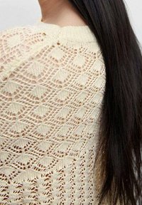 Pullover in crema con un motivo a traforato che presenta motivi a diamante e a foglia, scollatura rotonda e finitura testurizzata. Capelli lunghi e scuri visibili.