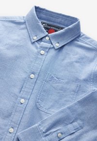 Vaaleansininen nappipaita, joka on valmistettu teksturoidusta kankaasta. Siinä on kaulus, valkoiset napit, rintatasku ja kirjailtu logo.