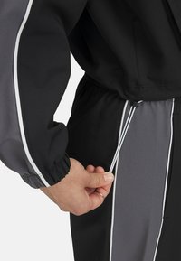 Veste de sport noire et grise avec des poignets élastiques ; présente des détails de piping blancs et un cordon de serrage ajusté à la taille.