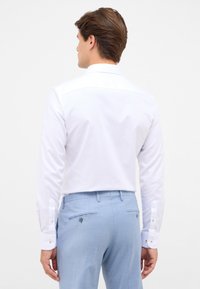 Weißes langärmliges Hemd mit glatter Textur und spitzen Kragen, kombiniert mit hellblauen Hosen mit Taschen auf der Rückseite und Knopfdetails.