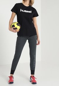 Camiseta negra con el logo "hummel", pantalones de chándal grises con el texto "mizuno", zapatillas deportivas rojas, sosteniendo una balón de mano amarillo, sobre un fondo claro.