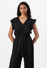 Combinaison noire avec décolleté en V, manches courtes à volants, boutons sur le devant et taille ceinturée. Fabriquée en tissu léger et texturé.