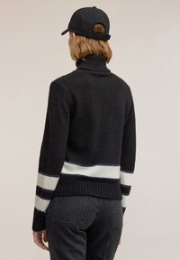 Czarny dziergany sweter z wysokim kołnierzem, zdobiony białymi i granatowymi paskami przy krawędzi i mankietach, zestawiony z czarnymi spodniami i czapką.