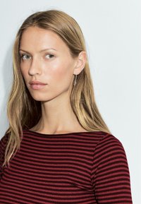 Woman with long, straight blonde hair wearing a burgundy and black striped ribbed top. Minimal silver earrings complement the look.