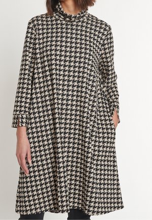 Frau trägt ein schwarz-beiges, langärmliges Kleid mit Hahnentrittmuster, hohem Kragen und Taschen, steht vor einem einfarbigen Hintergrund.