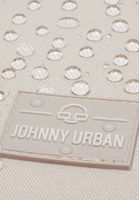 Közelről nézve bézs szövet vízcseppekkel és egy belevarrt "JOHNNY URBAN" címkével, amelyen egy minimalista arc logó látható.