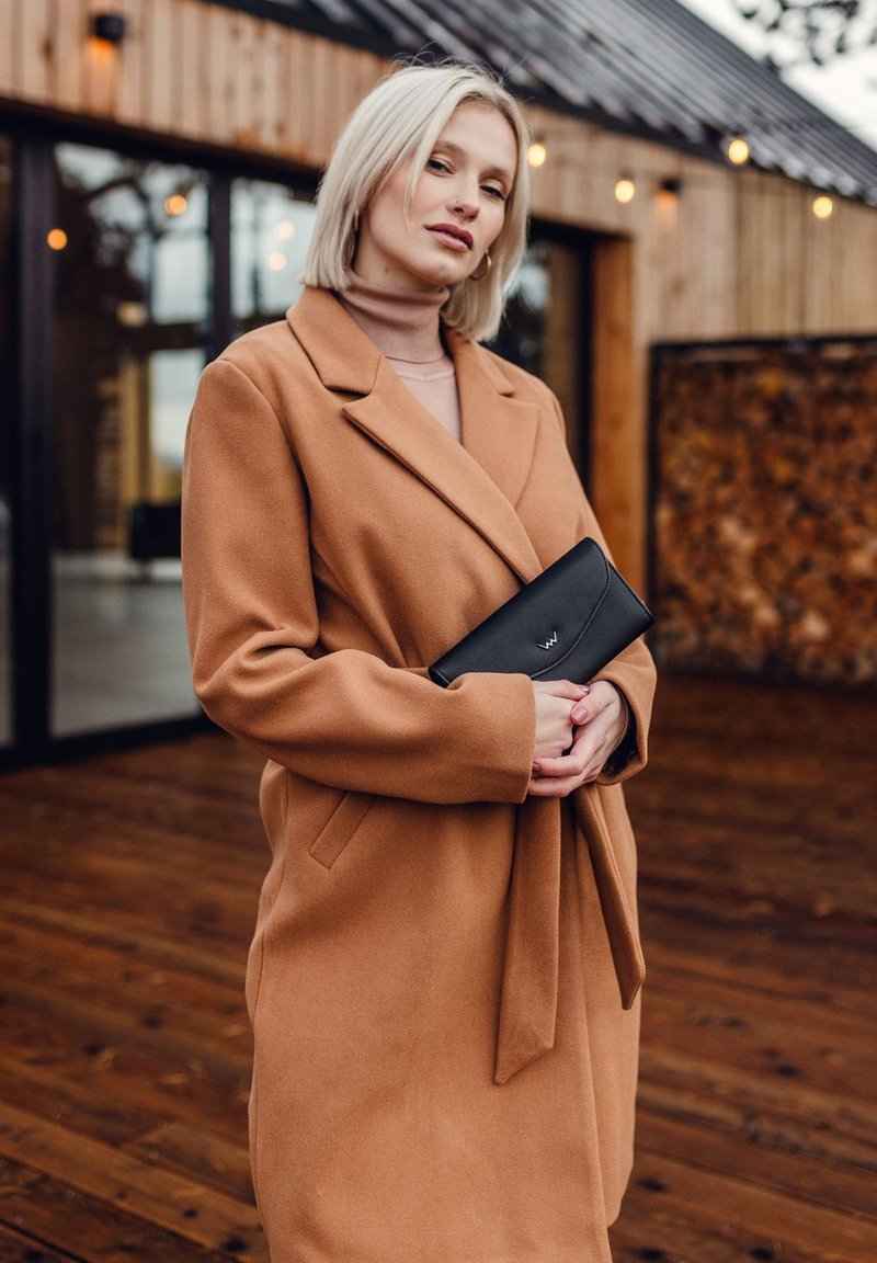 Béžový vlněný kabát se zářezovým límcem, bočními kapsami a opaskem. Drží černou kabelku s decentně embosovaným logem.