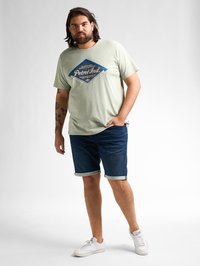 Lichtgroene t-shirt met korte mouwen met een diamantvormig blauw logo, gecombineerd met donkere denim shorts en witte sneakers. Eenvoudig ontwerp, casual pasvorm.