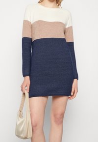 Robe en tricot présentant des rayures horizontales crème, beige et bleu marine. Manches longues et encolure ronde. Texture douce, longueur genou, assortie à un sac beige.