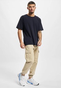 Jonge man met kort haar en tatoeages draagt een donkerblauw T-shirt, beige cargobroek en witte sneakers met blauwe accenten, staand op een witte achtergrond.