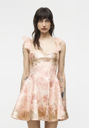 Sötét hajú nő, aki ujjatlan rózsaszín és arany virágmintás brokát ruhát visel masnis pántokkal, sima háttér előtt áll.