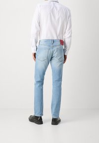 Světle modré džíny z denimu s úzkým střihem, vybavené zadními kapsami a červenou značkovou nášivkou v pase. Nošeny s bílým dlouhým rukávem.