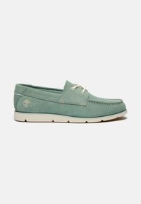 Scarpa da barca in suede verde con suola in gomma chiara, dettagli in pizzo e un piccolo logo di un albero sul lato. Design semplice e a profilo basso.