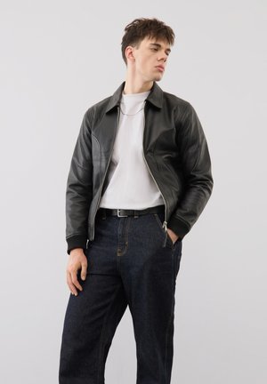 Junger Mann, der mit einer Hand in der Tasche steht, eine schwarze Lederjacke, ein weißes T-Shirt, dunkle Jeans und einen Gürtel vor einem einfarbigen Hintergrund trägt.
