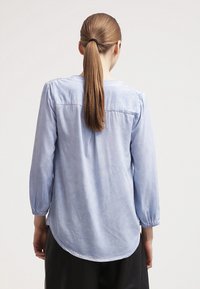 Kobieta z długimi jasnobrązowymi włosami związanymi w kucyk, ubrana w jasnoniebieską bluzkę z długim rękawem, stojąca na jednolitym tle.