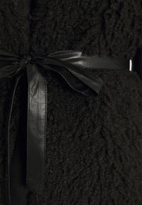 Czarna, teksturowana odzież wierzchnia o puszystym wykończeniu, z gładkim, czarnym skórzanym paskiem zawiązanym w talii, podkreślającym kształt odzieży.
