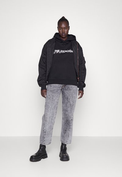 Black bomber jacket over a black hoodie with graphic text, paired with grey straight-leg jeans and chunky black boots. Minimalistic design.