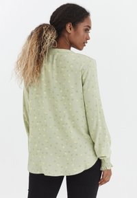 Lichtgroene blouse met lange mouwen, voorzien van bloemmotieven in geel en paars, elastische mouwen en een gebogen zoom.