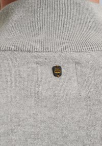 Hellgrauer Pullover mit einem gerippten Kragen und einem schwarzen Markenetikett mit goldener Schrift auf der Rückseite. Weiche, gestrickte Textur.