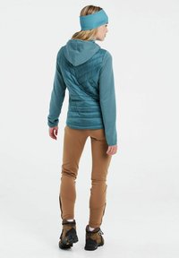 Mėlynas pūkų švarkas su raukšlėmis rankovėse, rudos lauko kelnės ir tamsūs kalnų batai. Lankelis atitinka švarko spalvą; elegantiškas, funkcionalus dizainas.