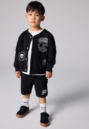 Jongentje staande, draagt een zwarte AllSaints jas en korte broek met witte grafische prints, witte AllSaints sokken en zwarte sneakers.