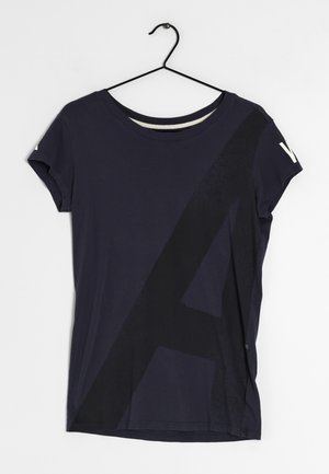 Mørkeblå T-shirt med korte ærmer og et stort mørkt geometrisk design på forsiden, hængende på en sort wirebøjle mod en hvid baggrund.