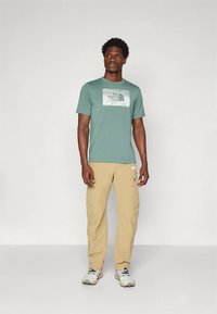 Meeste lühikeste varrukatega roheline t-särk graafilise logoga ees, kantud koos helebeeži pükste ja hallide tossudega. Lihtne disain, vaba lõige.