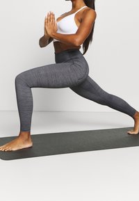Szare legginsy sportowe o teksturowanym wykończeniu, wysoki stan i bezszwowy design, w połączeniu z białym biustonoszem sportowym na ciemnej macie do jogi.